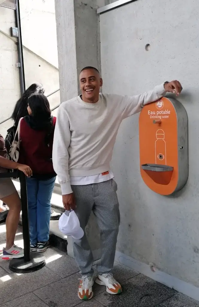 Jeune homme souriant à côté d'une fontaine à eau murale orange marquée "Eau potable" dans un espace public moderne.