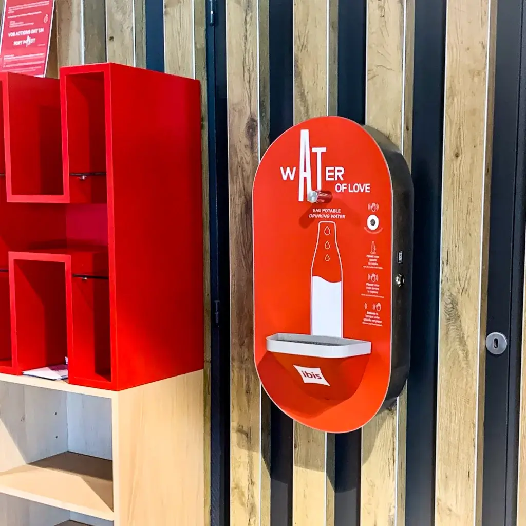 Fontaine à eau murale rouge "Water of Love" située dans un espace intérieur moderne avec un panneau en bois et des étagères rouges.