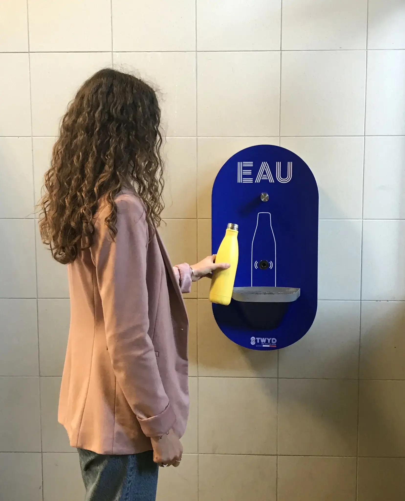 Femme utilisant une fontaine à eau Twyd avec une gourde jaune, symbole d'hydratation durable et écologique.